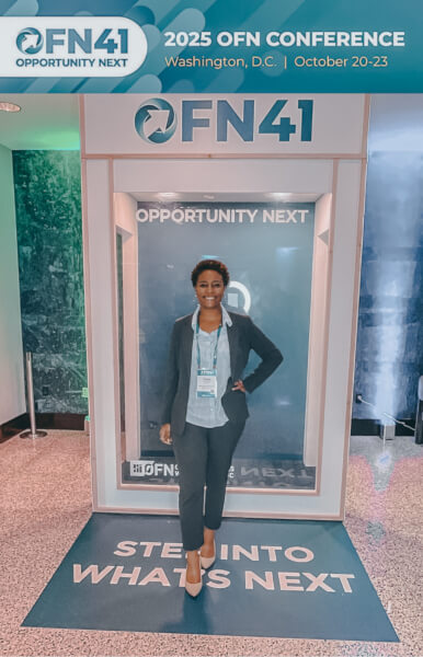Article author Casey Ariel Dike’ posing at the OFN Conference in front of a branded “OFN41 – Opportunity Next” banner.